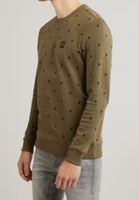 Felpa verde oliva con motivi a quadrati neri, collo e polsini a coste, con una piccola toppa logo sul petto.