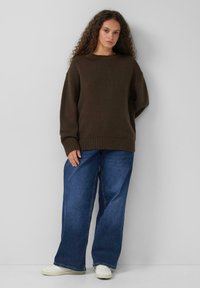 Donkerbruin gebreide trui met een ronde halslijn en geribbelde mouwen, gecombineerd met brede blauwe jeans en witte schoenen.