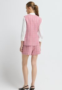 Růžová souprava z tkaného materiálu, vesta a šortky, s texturovanou látkou a bílou košilí pod ní, doplněná černými otevřenými podpatky. Zobrazeno ze zadní strany.