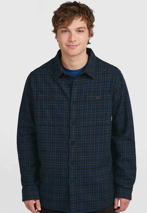 Junger Mann trägt ein blau-schwarz kariertes Hemd über einem dunkelblauen T-Shirt und steht vor einem schlichten hellen Hintergrund.