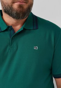 Zielona koszulka polo z teksturowanej tkaniny, z ciemnoniebieskimi paskami na kołnierzyku i małą naszywką z logo na klatce piersiowej.