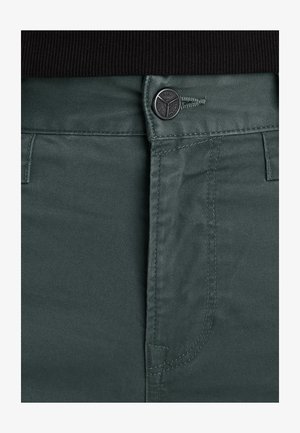 Close-up van de taille van groene broek met een zwarte knoop waarop 'PME Legend Jeans' staat, en zichtbare stiksel details op de stof.