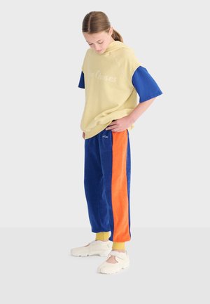 Jeune personne debout, portant un sweat à capuche jaune avec des manches bleues, un pantalon bleu et orange, et des chaussures blanches sans lacets, regardant son tenue.