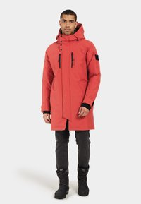 Röd vattentät parkas med huva, med svarta dragkedjor och detaljer, två framfickor och knälång design. Buren över mörka jeans och stövlar.