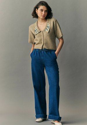 Cardigan en tricot beige à manches courtes et broderie florale sur le col, associé à un pantalon en denim bleu ample et des chaussures blanches.