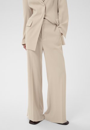 Bézs, széles szárú nadrág egy hozzá illő bézs blézerrel párosítva, fekete, négyzet alakú orrú cipőkkel, álló modellen bemutatva.