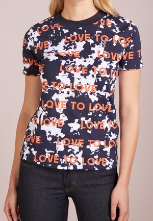 Donna che indossa una maglietta a maniche corte blu navy e bianca con un motivo e testo arancione che ripete "LOVE TO LOVE", abbinata a jeans scuri.