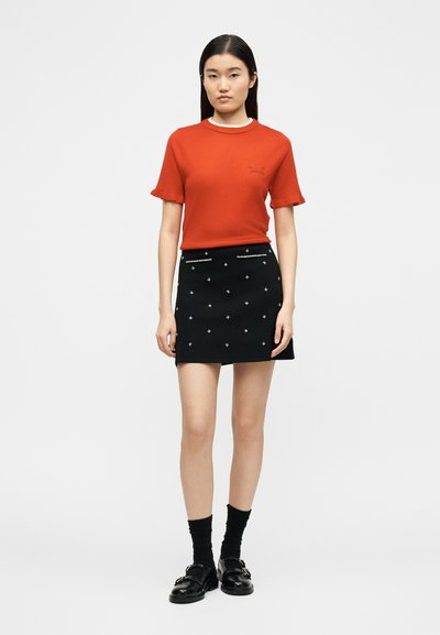 Νέα γυναίκα που φοράει κόκκινο κοντομάνικο τοπ και μαύρη μίνι φούστα με σχέδιο λευκές τελείες, μαύρες κάλτσες και μαύρα μοκασίνια, στέκεται μπροστά σε λευκό φόντο.