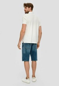 Mann trägt ein weißes kurzärmliges Polohemd, blaue Jeans-Shorts in Kniehöhe und weiße Sneaker, steht und schaut über seine Schulter.