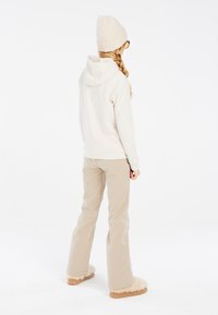 Felpa crema con una tasca sul retro, pantaloni beige a zampa d'elefante e pantofole morbide color nocciola. Un berretto di maglia con un motivo testurizzato completa l'outfit.