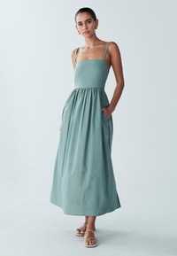 Rento, puolipitkä mekko pehmeästä vihreästä kankaasta, jossa on istuva yläosa, kerätty vyötärö ja sivutaskut. Minimalistinen muotoilu, sileä rakenne.