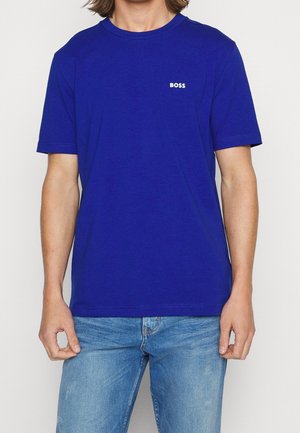 Person trägt ein schlichtes, königsblaues T-Shirt mit kurzen Ärmeln und dem "BOSS"-Logo auf der Brust sowie hellblaue Jeans.