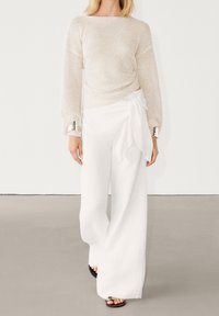 Hvide bukser med vide ben kombineret med en cremefarvet mesh-langærmet top med sølvdetaljer på manchetterne, der skaber et afslappet look.