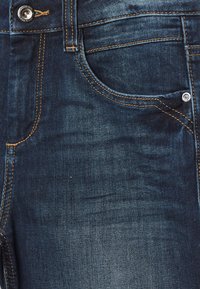 Primer plano de unos pantalones de mezclilla azul oscuro que muestran el bolsillo delantero, un pequeño bolsillo para monedas, costuras naranjas, y detalles de botón y remaches metálicos.