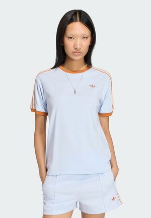 Kobieta w jasnoniebieskich sportowych szortach Adidas i koszulce z pomarańczowymi lamówkami i logo, stojąca z rękami w kieszeniach na jednolitym tle.