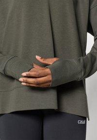 Camiseta de manga larga verde oliva con agujeros para los pulgares, que presenta un diseño relajado, textura suave y detalles de costura visibles. Leggings negros por debajo.