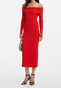 Robe rouge à côtes, épaules dénudées, manches longues, silhouette ajustée, et talons noirs à bride cheville. Accessoires incluant un sac à main à motifs.