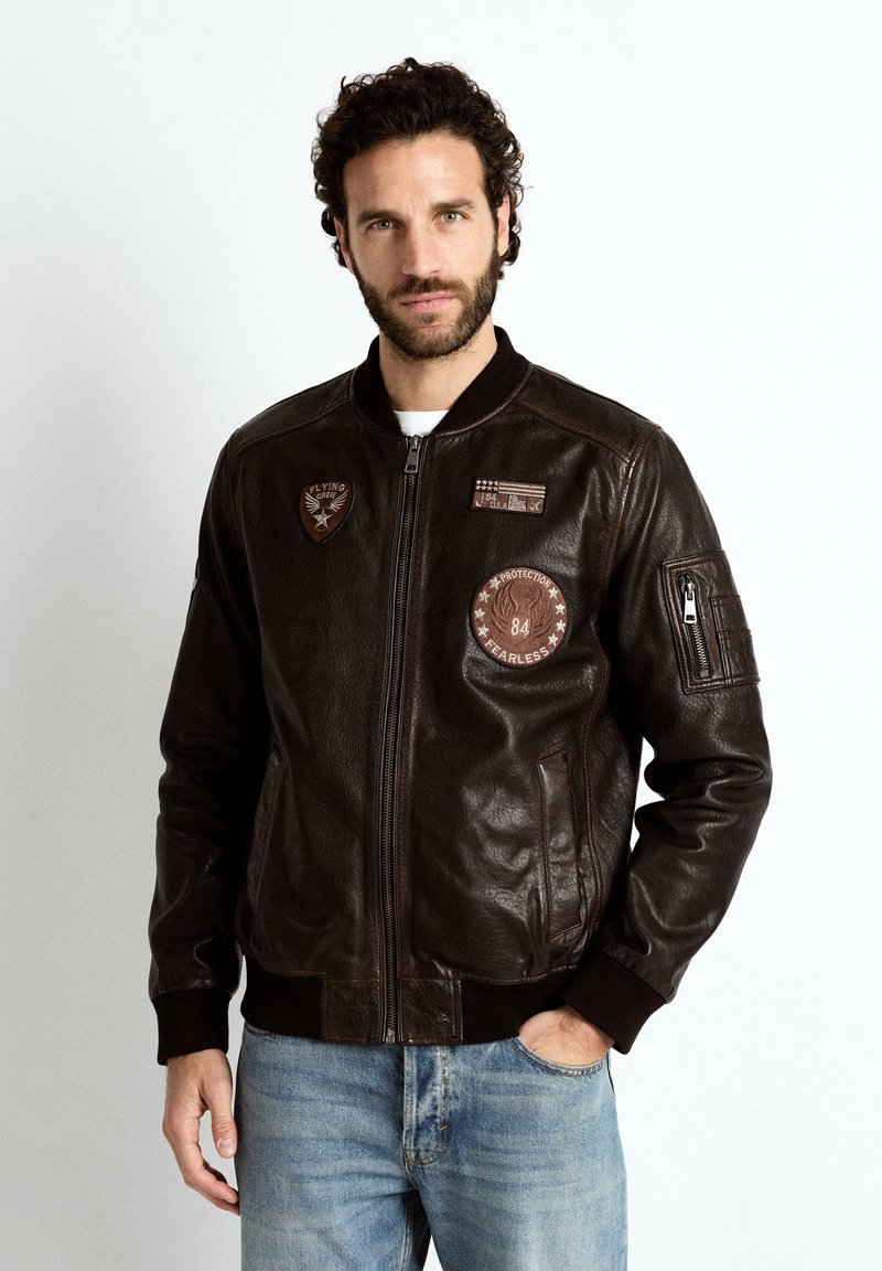 Mann mit dunklem lockigem Haar und Bart, der eine braune Leder-Bomberjacke mit Aufnähern und hellblaue Jeans vor einem weißen Hintergrund trägt.