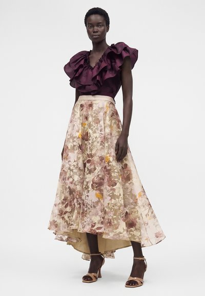 Γυναίκα που φοράει σκούρο μοβ τοπ με βολάν σε συνδυασμό με φούστα σε μπεζ χρώμα με λουλουδένιο μοτίβο και nude πέδιλα με τακούνια, στέκεται με φόντο λευκό.