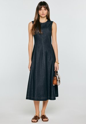 Kobieta w ciemnej jeansowej sukience midi bez rękawów, trzymająca małą dwukolorową torebkę, nosi brązowe płaskie sandały i złotą bransoletkę.