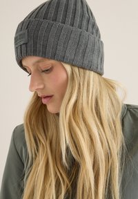 Szara, prążkowana czapka beanie z podwiniętym brzegiem i prostokątną łatą z logo. Wyróżniająca się tekstura kontrastuje z gładkim, neutralnym tłem.