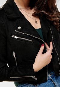 Zwart suède gecropte jas met zilveren ritsen, voorzien van meerdere ritssluitingen en een puntige kraag. Zachte textuur en gestructureerd ontwerp.