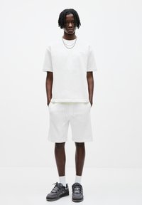 T-shirt en coton blanc et shorts assortis, tous deux avec une coupe décontractée. Finition avec un branding subtil, accompagnés de chaussures de sport noires.