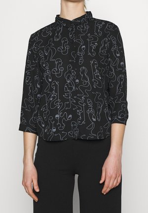 Chemise noire à boutons avec des dessins en ligne continue blanche de visages humains abstraits et de formes, portée avec un pantalon noir.