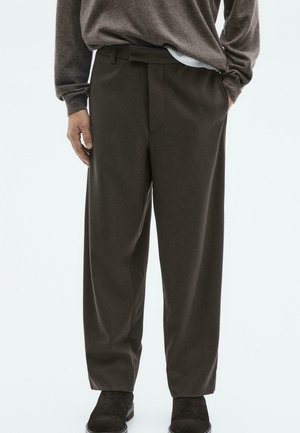 Person wearing dark brown wide-leg trousers, brown sweater, white undershirt, and black suede ankle boots, with hands partially in pockets.