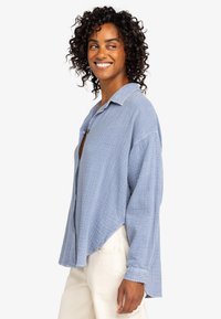 Light blue, textured, long-sleeve shirt with a buttoned collar and a relaxed fit, featuring a curved hem and drop shoulders.