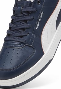 Gros plan d'une basket en cuir bleu marine et blanc avec un marquage "Porsche" et des lacets bleu marine sur une semelle blanche.