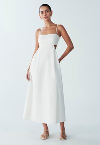 Vestido maxi de lino blanco con escote cuadrado, tirantes finos y cortes laterales; presenta una falda fluida y una tela con textura sutil.