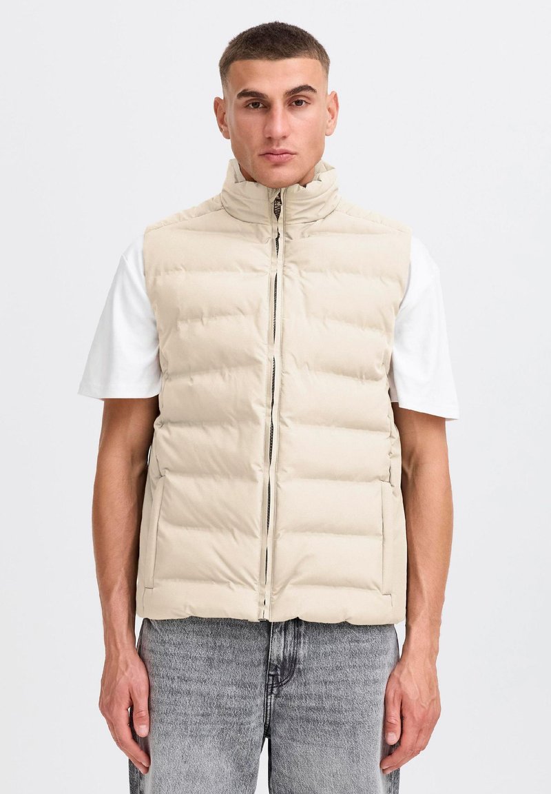 Homme debout devant un fond uni, portant une doudoune beige sur un t-shirt blanc et un jean gris, face à l'avant.