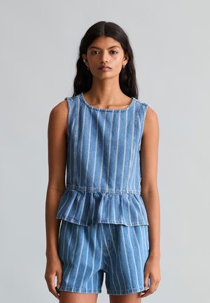 Frau mit langen dunklen Haaren, die ein ärmelloses blau-weiß gestreiftes Denim-Top mit Peplum und passende Shorts trägt, steht vor einem grauen Hintergrund.