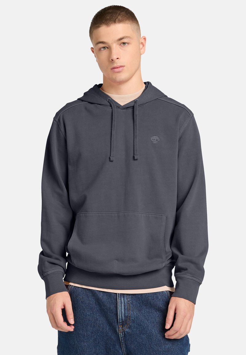 Joven vistiendo una sudadera con capucha gris oscuro con bolsillo frontal y un sutil logo de árbol, combinada con jeans azules sobre un fondo claro.
