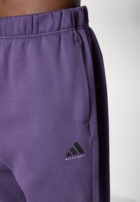 Fialové tepláky s hladkou texturou, elastickým pasem a postranními kapsami. Obsahují černé logo Adidas s textem "BASKETBALL".