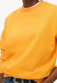 Sudadera amarilla con cuello redondo, puños y dobladillo acanalados. Textura suave y lisa; combinada con un cinturón negro y jeans de mezclilla.