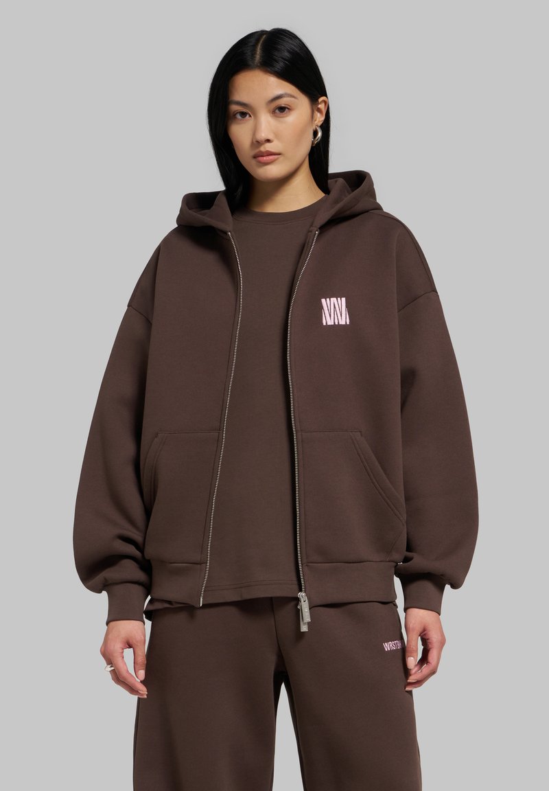 Νέα γυναίκα που φοράει καφέ oversized φούτερ με φερμουάρ και matching φόρμα, στέκεται μπροστά σε απλό γκρι φόντο.