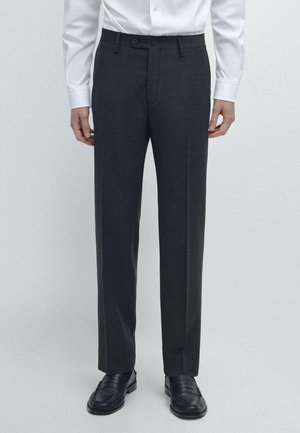 Man die donkergrijze op maat gemaakte pantalon draagt, zwarte leren loafers en een wit, met knopen gesloten overhemd, staand tegen een lichte effen achtergrond.