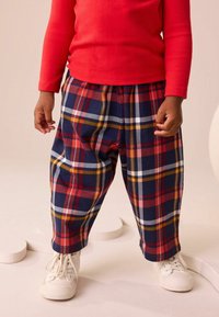 Cămașă roșie cu mâneci lungi, asociată cu pantaloni lejeri, din material șcheckered în nuanțe de bleumarin, roșu, galben și alb. Copilul poartă adidași albi.