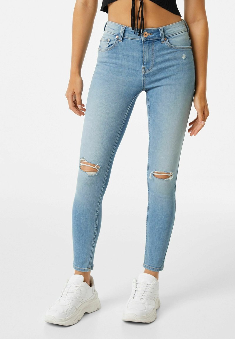 Vaqueros skinny de color azul claro con dos detalles de rodillas rasgadas, cinco bolsillos y cierre de botón. Combinados con zapatillas blancas. Textura de denim suave.