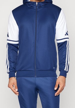 Homme portant un sweat à capuche zippé bleu marine et un pantalon assorti avec des manches rayées blanches et bleues marine, debout les mains détendues.