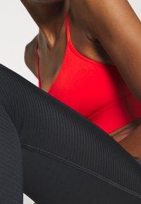 Červená sportovní podprsenka s tenkými ramínky ve spojení s černými žebrovanými legínami. Látka je pružná a těsně přiléhá. Jsou vidět detaily švů.