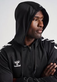 Felpa nera con zip realizzata in un tessuto morbido, dotata di cappuccio, logo bianco e accenti bianchi sui lati con motivo a zigzag sulle maniche.