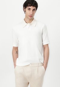 Hvit kortermet polo skjorte med teksturert stoff og en kontrasterende beige krage. Kombinert med lysebeige mønstrede bukser med en knapp.