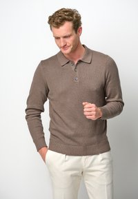 Brauner gestrickter Pullover mit einem klassischen Kragen und einer Knopfleiste mit drei Knöpfen. Rippbündchen und Saum; glatte Textur. Über hellen Hosen getragen.