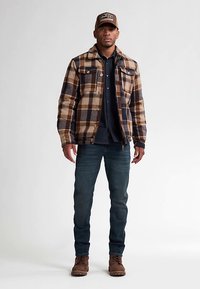 Geruite jas in bruin en marineblauw, met twee borstzakken en knopen, gedragen over een donker shirt. Blauwe spijkerbroek en bruine laarzen maken de outfit compleet.