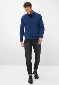 Maglione blu con zip al collo in maglia texture, abbinato a jeans neri con maniche arrotolate e stivali neri con lacci. Design semplice, silhouette aderente.