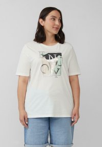 Frau mit dunklen Haaren, die ein weißes Grafik-T-Shirt und helle blaue Jeansshorts trägt, schaut seitlich vor einem schlichten Hintergrund.