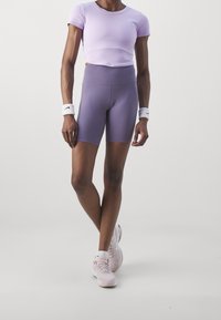 Jasnofioletowy crop top z okrągłym dekoltem, spodenki z wysokim stanem w jasnofioletowym kolorze oraz różowe buty sportowe. Podkreślone białymi opaskami na nadgarstki.